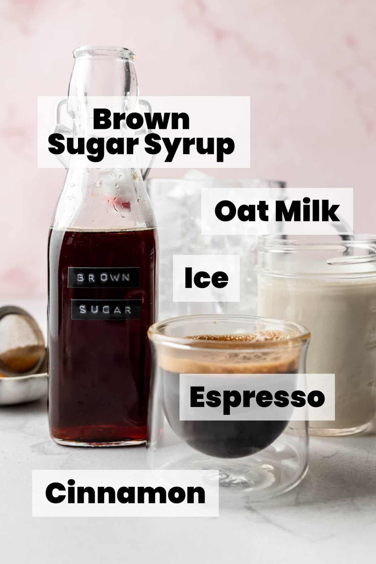 A bottle of homemade brown sugar syrup labeled “BROWN SUGAR” sits on a white surface alongside a glass of espresso, a jar of milk, and a pitcher of ice, with a soft pink marbled background.