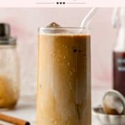 A cold brown sugar shaken espresso in a tall glass with a glass straw, condensation forming on the outside. In the background, a bottle of brown sugar syrup, a shaker, and scattered ingredients are visible.