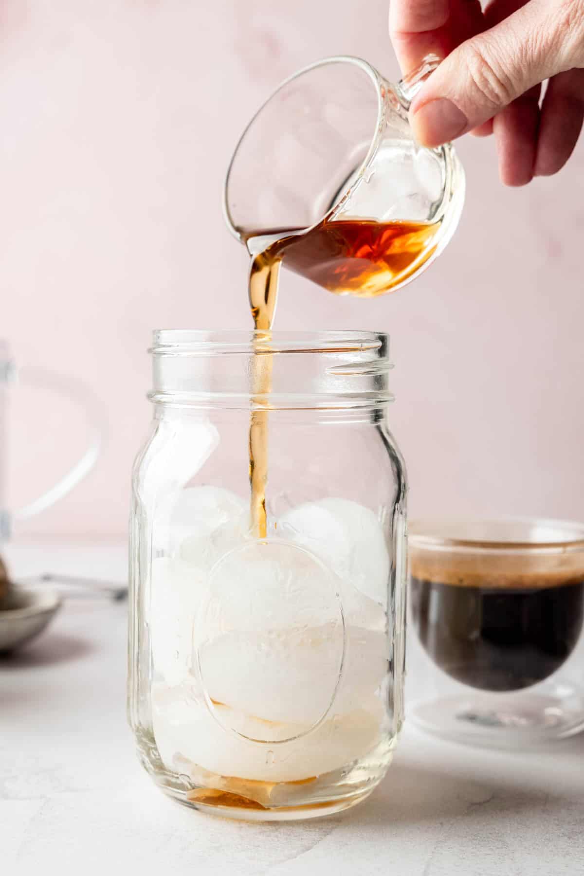 A hand pours brown sugar syrup into a mason jar filled with ice, with a double-walled glass of espresso sitting nearby.