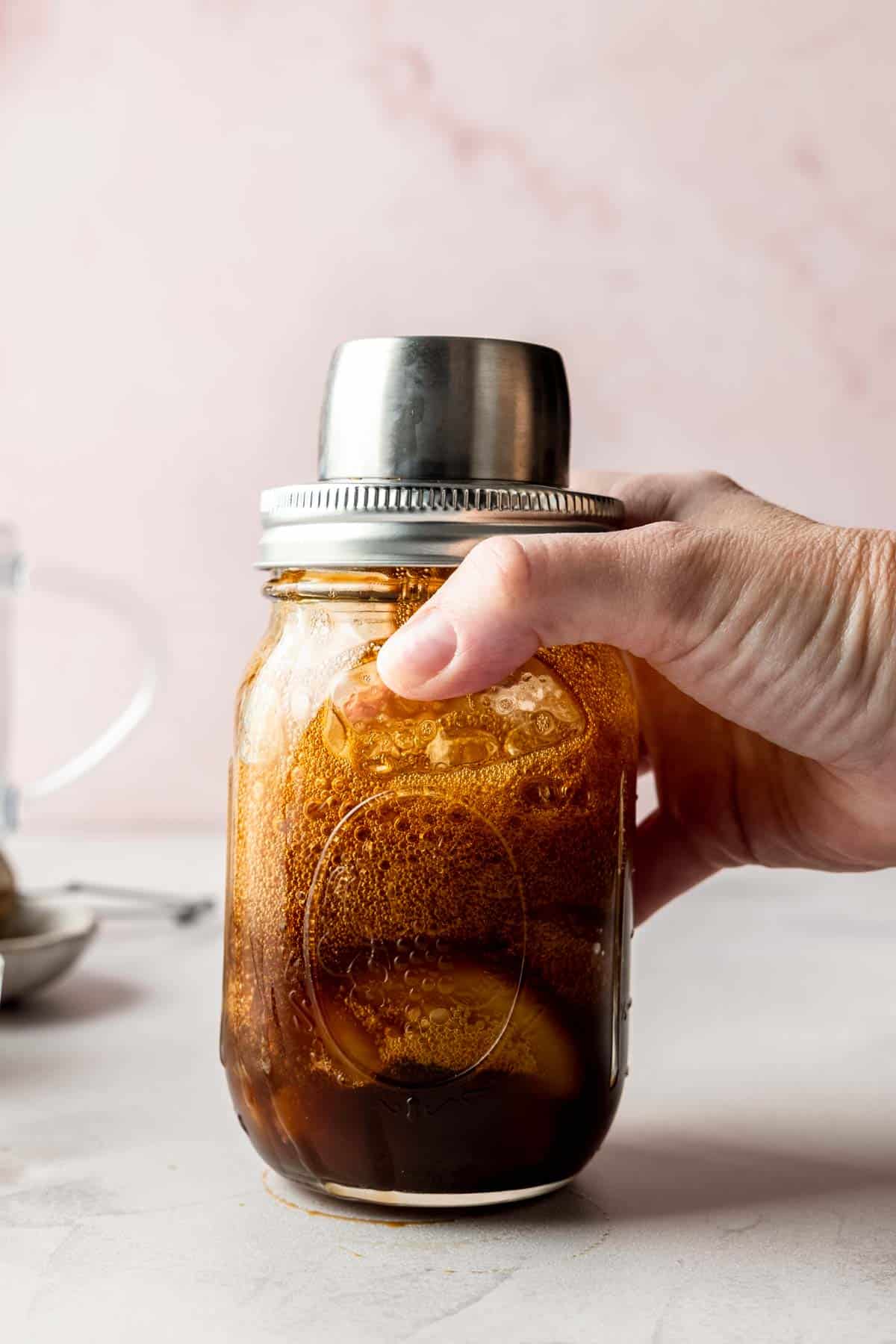A hand secures a cocktail shaker lid on a mason jar filled with espresso, ice, and syrup, preparing to shake.