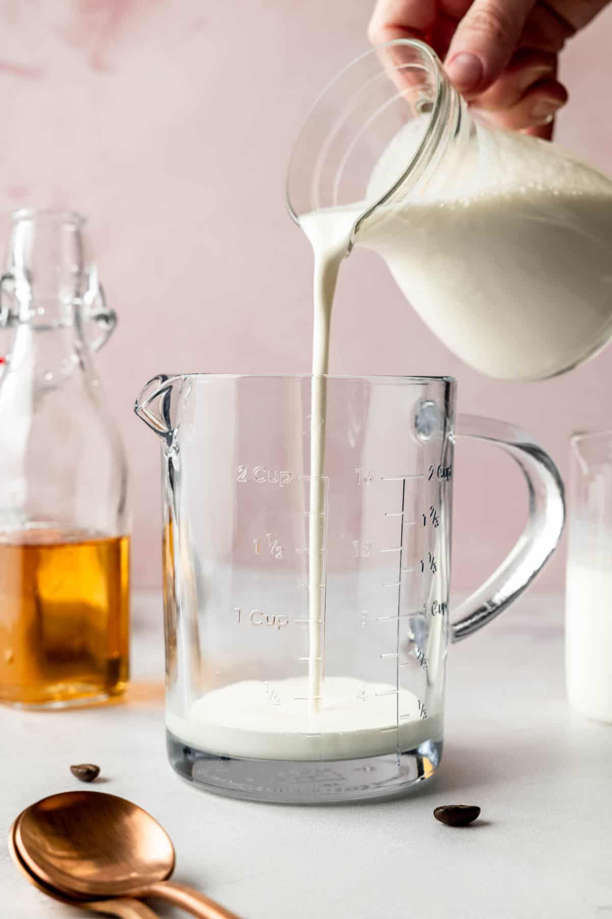 Milk being poured into a glass measuring cup, with a bottle of vanilla syrup and copper spoons in the background.