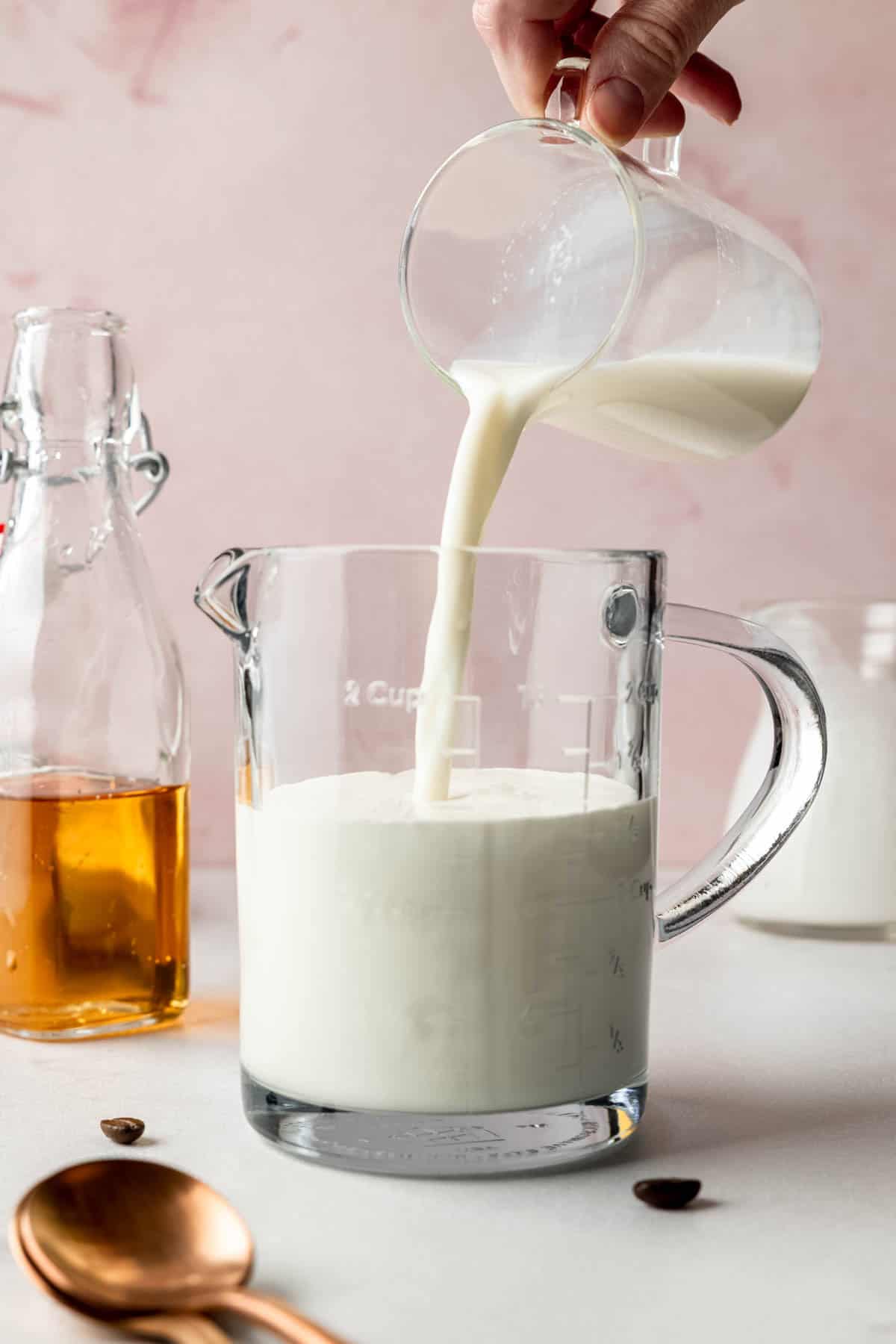 Additional milk being poured into the measuring cup, nearly filling it, with vanilla syrup visible behind.