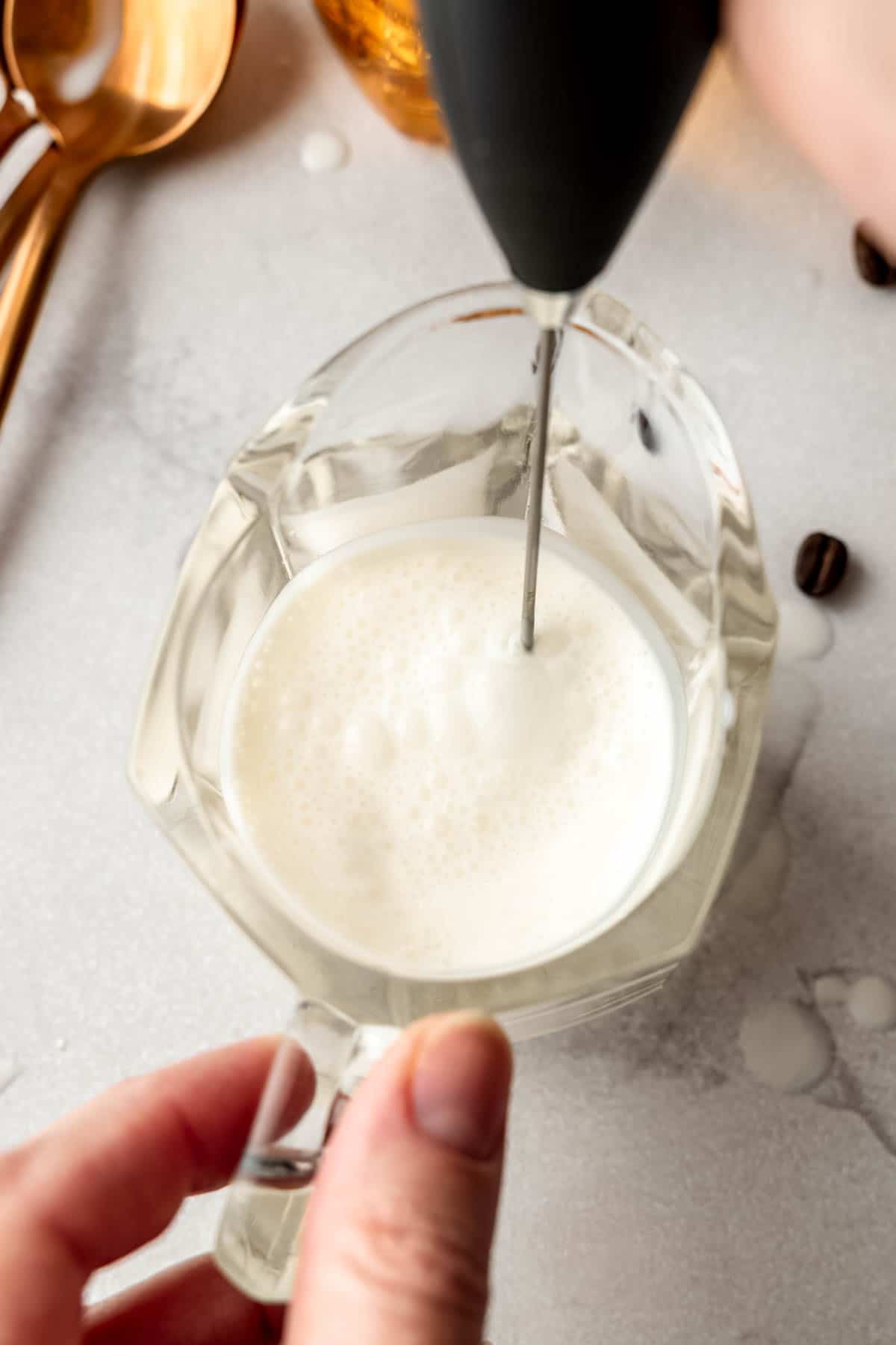 Close-up of a handheld milk frother whipping vanilla sweet cream in a glass measuring cup, beginning to foam.