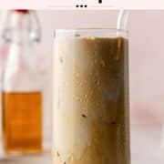 Final glass of vanilla shaken espresso with ice and a glass straw, condensation forming on the outside, with a bottle of vanilla syrup in the background.