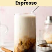 A tall glass of brown sugar shaken espresso with creamy milk swirls, served with a glass straw. Cinnamon sticks, coffee beans, and brown sugar crystals are scattered around on a light surface.