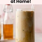 Final glass of vanilla shaken espresso with ice and a glass straw, condensation forming on the outside, with a bottle of vanilla syrup in the background.