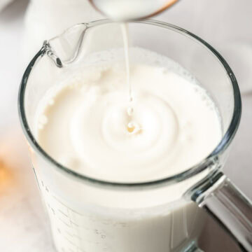 A spoon lifted above the prepared sweet cream mixture, with a drop falling back into the glass measuring cup.
