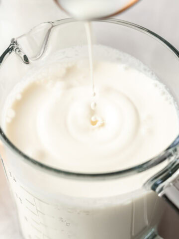 A spoon lifted above the prepared sweet cream mixture, with a drop falling back into the glass measuring cup.