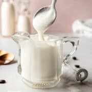 A spoonful of vanilla sweet cream cold foam held above a glass pitcher filled with freshly frothed foam.