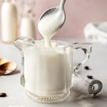 A spoonful of vanilla sweet cream cold foam held above a glass pitcher filled with freshly frothed foam.
