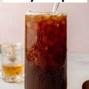 Finished glass of coffee soda with a straw, surrounded by coffee beans, syrup, and espresso in the background.