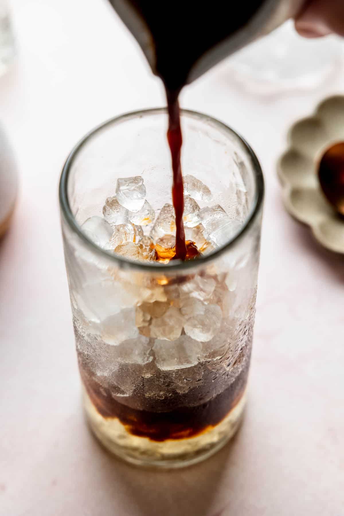 Pouring espresso over ice and syrup in tall glass.