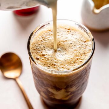 Pouring milk into a tall glass filled with frothy shaken espresso.