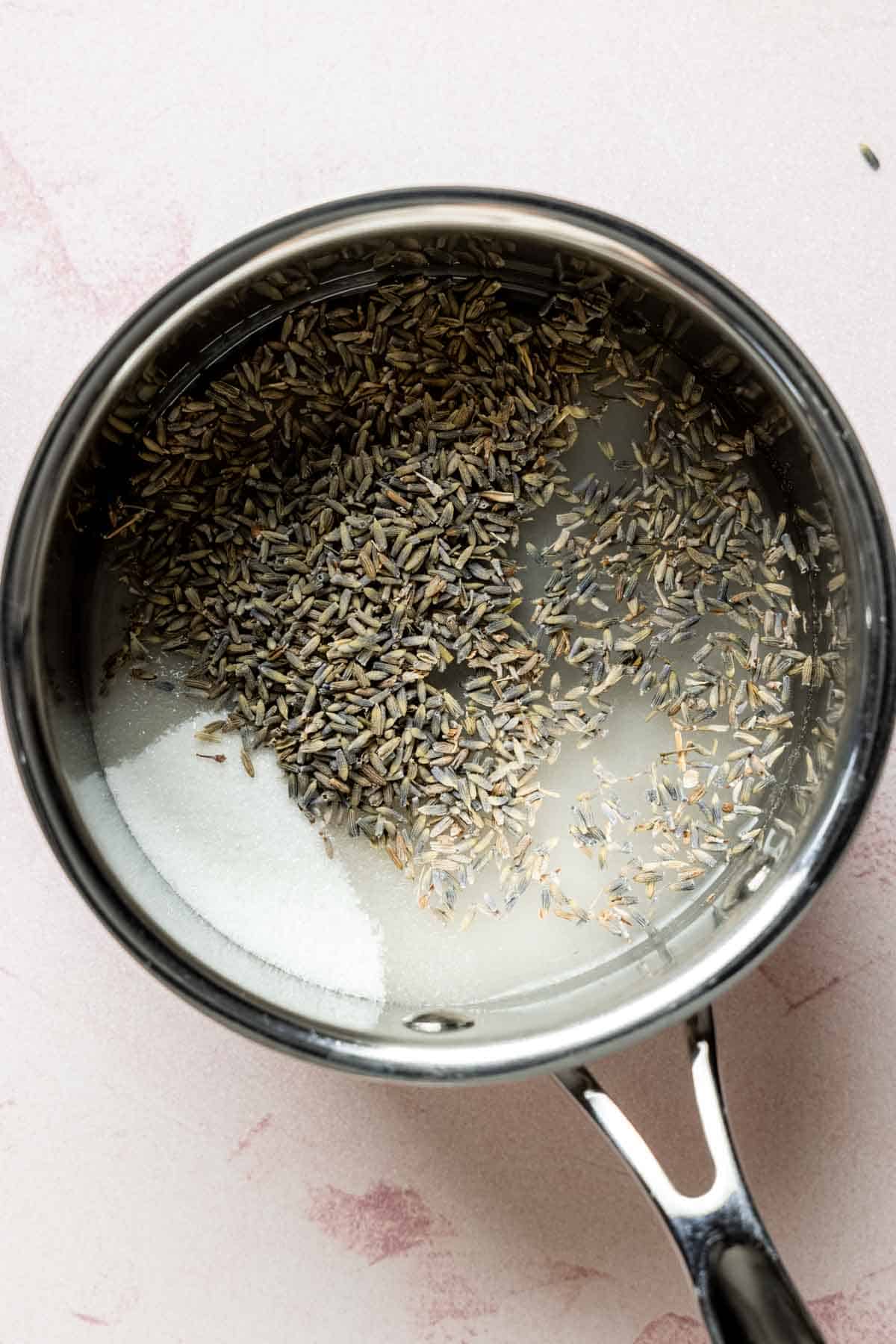 A pan filled with lavender buds, sugar, and water on a pink background.