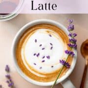 A lavender latte in a mug with foam, lavender buds, and a sprig of fresh lavender resting on top.